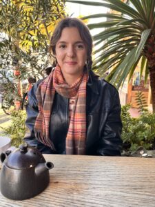 Lucia sitting at an outside table wearing a leather jacket and a tartan scarf smiling.